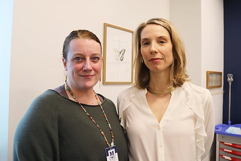 Two women standing indoors, one wearing a green top with a lanyard, the other in a white blouse; framed artwork and medical equipment are visible in the background.
