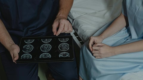A doctor in blue scrubs shows a patient an X-ray image, pointing at it while seated on a hospital bed.