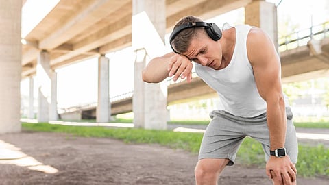 A tired man in workout clothes rests under a bridge after exercising, wearing headphones.