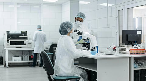 Scientists wearing protective gear work in a lab. One uses a microscope, while another stands nearby. 
