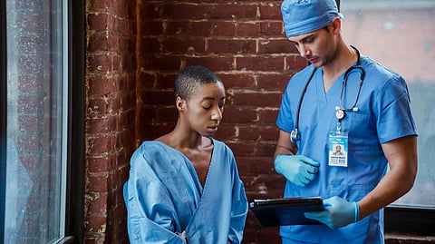 A nurse in blue scrubs and cap talks to a patient in a blue gown, holding a tablet.