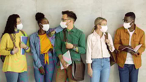 Five diverse students wearing face masks and casual clothes stand against a wall, holding books and bags.