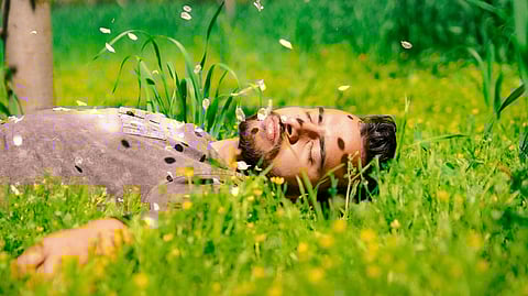 Man lying in grass surrounded by yellow wildflowers with white flower petals floating in the air during pollen season 
