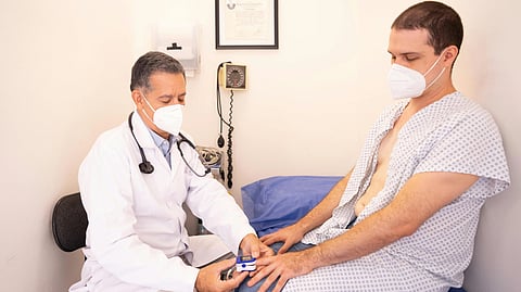 A doctor wearing a white coat and mask checks a patient's vitals using a pulse oximeter.