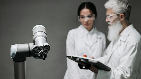 A robotic arm is in the foreground, with two scientists in lab coats and safety glasses in the background.