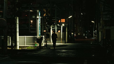 A person walks alone on a dimly lit urban street at night, surrounded by streetlights and faint building outlines.