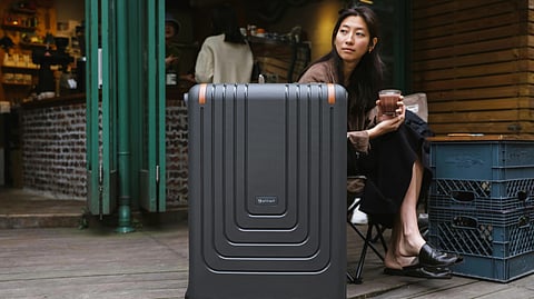 A woman is sitting outside a café, holding a drink and looking to the right. A black suitcase is placed in front of her.