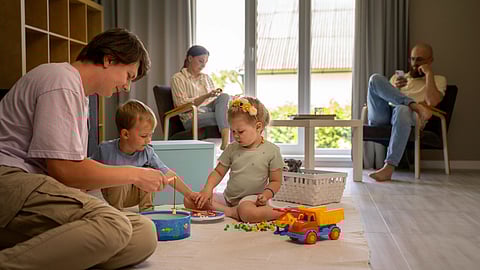A family in a cozy living room: two children playing with toys on a rug, an adult joining them.