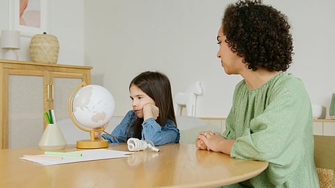 A woman and a young girl sit at a table with a globe, papers, and pencils.