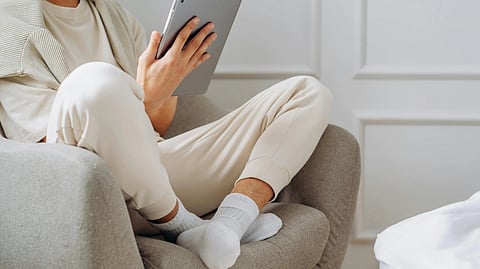 A person in a cozy sweater and sweatpants sits in an armchair, holding a tablet. 