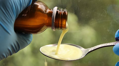 A person in blue gloves pours a thick, yellow liquid from a brown bottle onto a silver spoon.