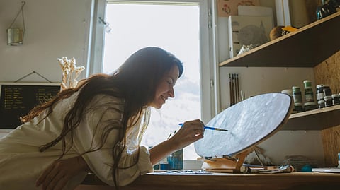 A woman smiling while painting on a canvas in a sunlit art studio with art supplies in the background.