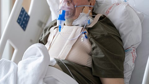 An older man, a patient in intensive care, lies in a hospital bed wearing an oxygen mask and chest binder, surrounded by medical monitors and supported by pillows.