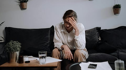 A woman in a white shirt sits on a dark sofa, looking stressed with a hand on their forehead. 