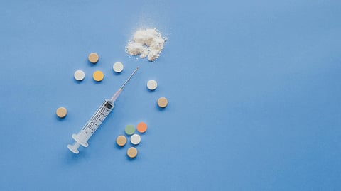 A syringe, assorted colorful pills, and a small mound of white powder are arranged on a blue background.