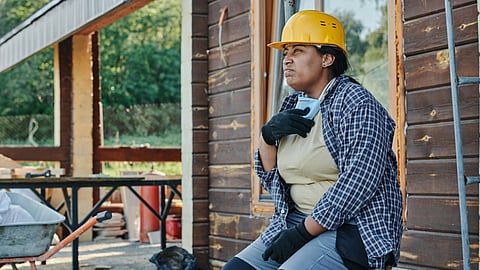 A construction worker in a yellow hard hat sits thoughtfully against a wooden building.