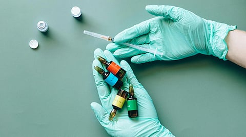 Gloved hands holding colorful vials and a syringe over a green background.