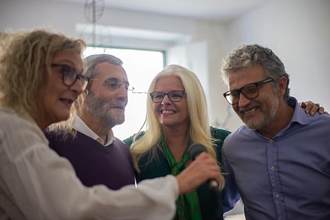 Four smiling adults, two women and two men, stand closely together indoors. One woman holds a microphone.