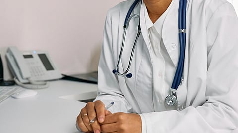 A doctor in a white coat with a stethoscope sits at a desk, holding a pen over a document and using a phone.