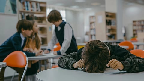 A child rests their head on a table, looking upset, while three other kids talk in the background.