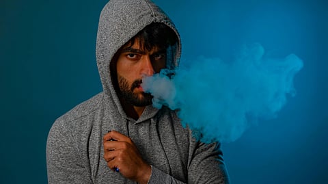 Man in a gray hoodie exhaling blue smoke from vaping against a cool-toned blue background.
