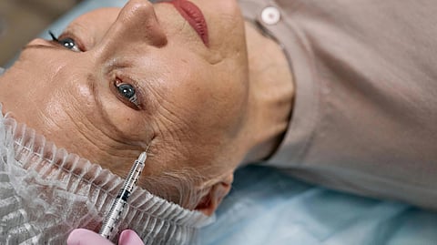 An elderly woman receives a cosmetic injection near her eye. She lies on a medical table wearing a cap. 