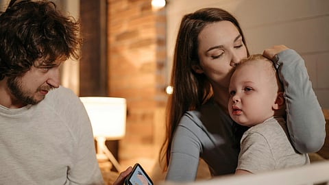 A cozy family scene in a softly lit room. A woman gently kisses a baby's head.