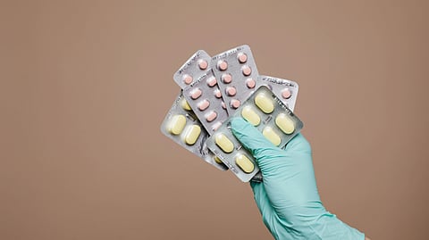 A hand in a teal glove holds blister packs of pink and yellow pills against a plain beige background.
