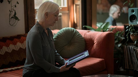 A white-haired woman sits on an orange sofa holding a frame in warm sunlight.