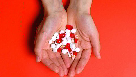 Hands hold a variety of red and white pills against a vibrant red background.