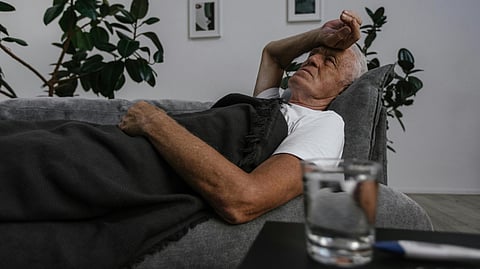 Elderly man resting on a couch, covered with a dark blanket, hand on forehead.