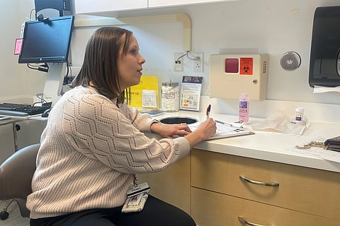 A woman sitting at a desk in a hospital room, focused on her work with medical equipment in the background.