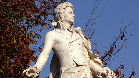 A white marble statue of a man in 18th-century attire stands elegantly against an autumn background. 