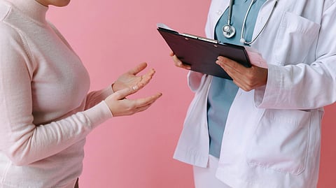 A patient in a pink sweater gestures while talking to a doctor in a white coat holding a clipboard. 