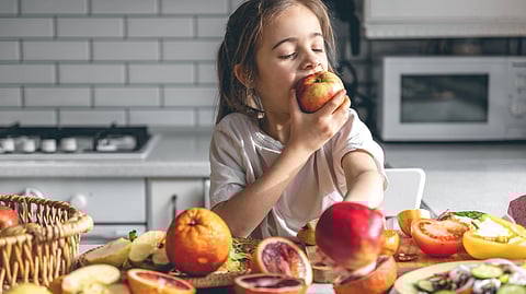 A little girl is eating an apple from a table filled up with fruits. 
