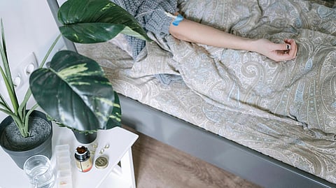 A person lies in bed under a patterned blanket with an IV in their arm. 