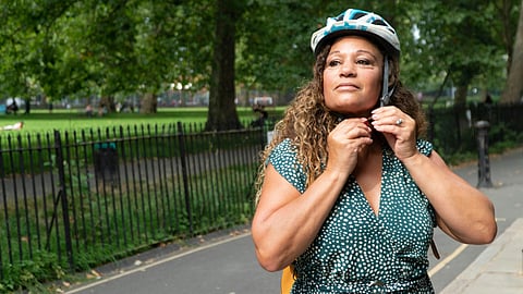 A woman in a green polka dot dress secures her helmet strap while standing on a path in a park. 