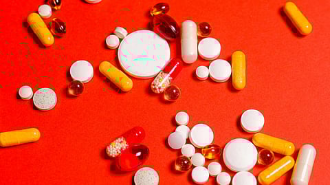 Various pills and capsules in white, yellow, red, and transparent colors are scattered on a vibrant red background.