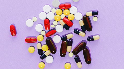 A variety of colorful pills and capsules, including red, yellow, and white, scattered on a light purple background.
