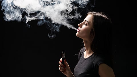Young beautiful brunette woman smoking, vaping e-cigarette with smoke on the black wall.