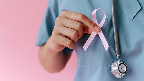 A female healthcare worker in scrubs holds a pink ribbon, symbolizing breast cancer awareness, against a pink background.