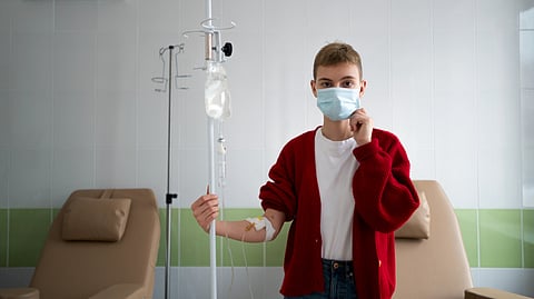 A person wearing a red cardigan and face mask in a medical room holds an IV stand.