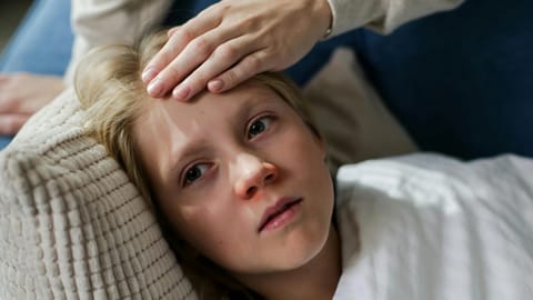 A concerned hand feels a young person's forehead as they lie on a pillow, wrapped in a blanket. 