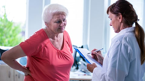 An elderly woman in a red shirt, with a pained expression, holds her lower back. A doctor in a white coat attentively takes notes on a clipboard. 
