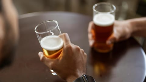 Two people hold glasses of beer over a dark round table. 