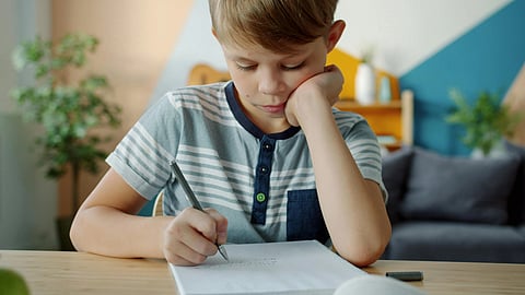 A young boy with short hair focuses intently on writing in a notebook at a wooden table.