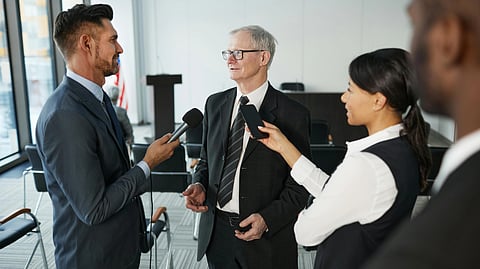 A formal setting with a senior man in a suit being interviewed by two journalists holding microphones. 