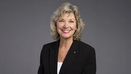 Smiling Kim Orth with short blond hair wearing a black blazer over a white top against a gray background.