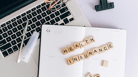 Laptop planner with “Health Insurance” spelled in tiles, with pen, glasses, and clip nearby.