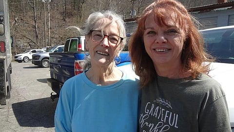 Two women smiling outdoors in a sunny parking lot, surrounded by cars. 
